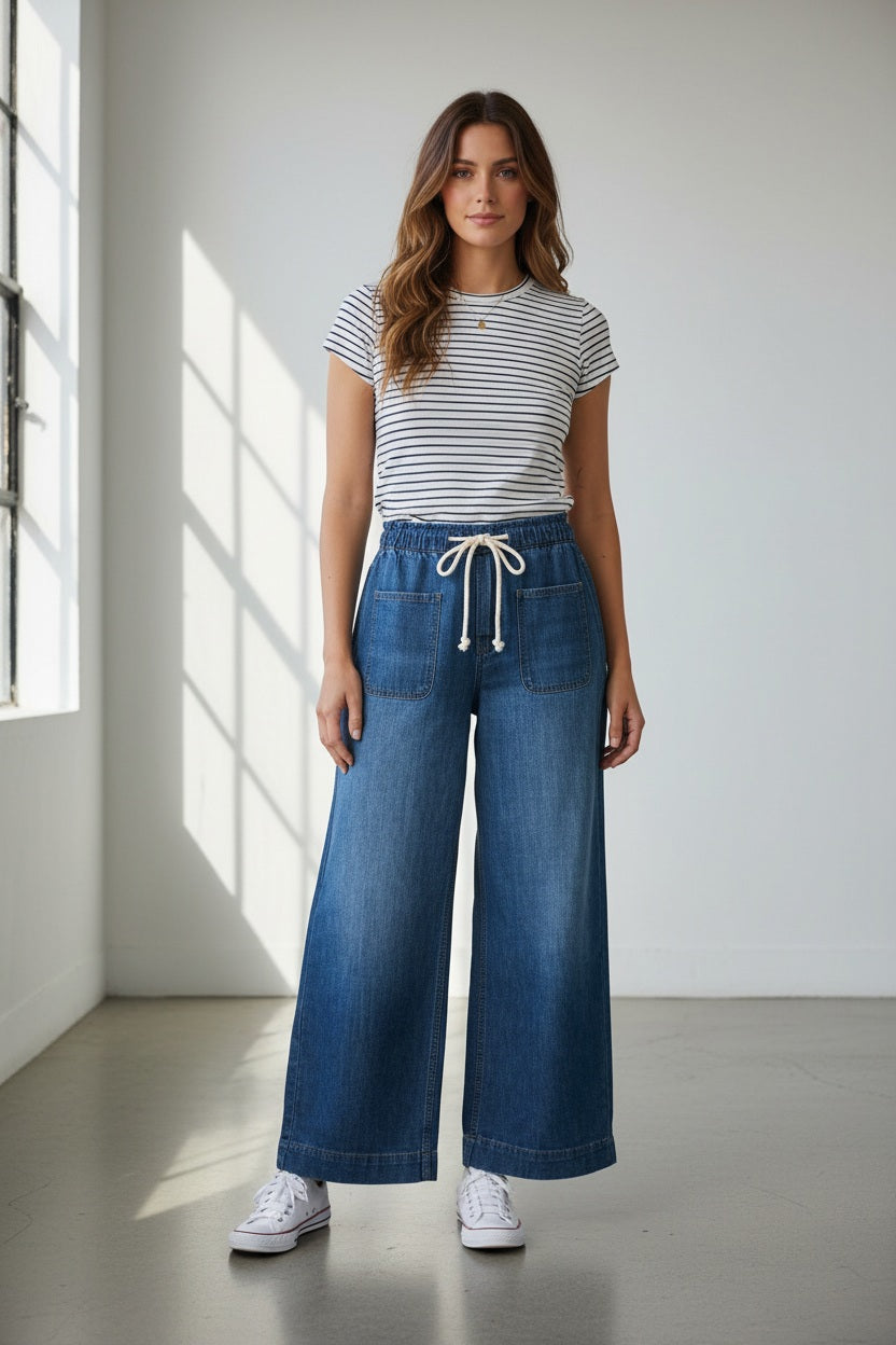Wearing MERSEA's LONDONBERRY Drawstring Supersoft Denim Pant with wide legs and large front pockets, paired with a light grey top, gold jewelry, and brown sandals, a person poses against a plain light background.