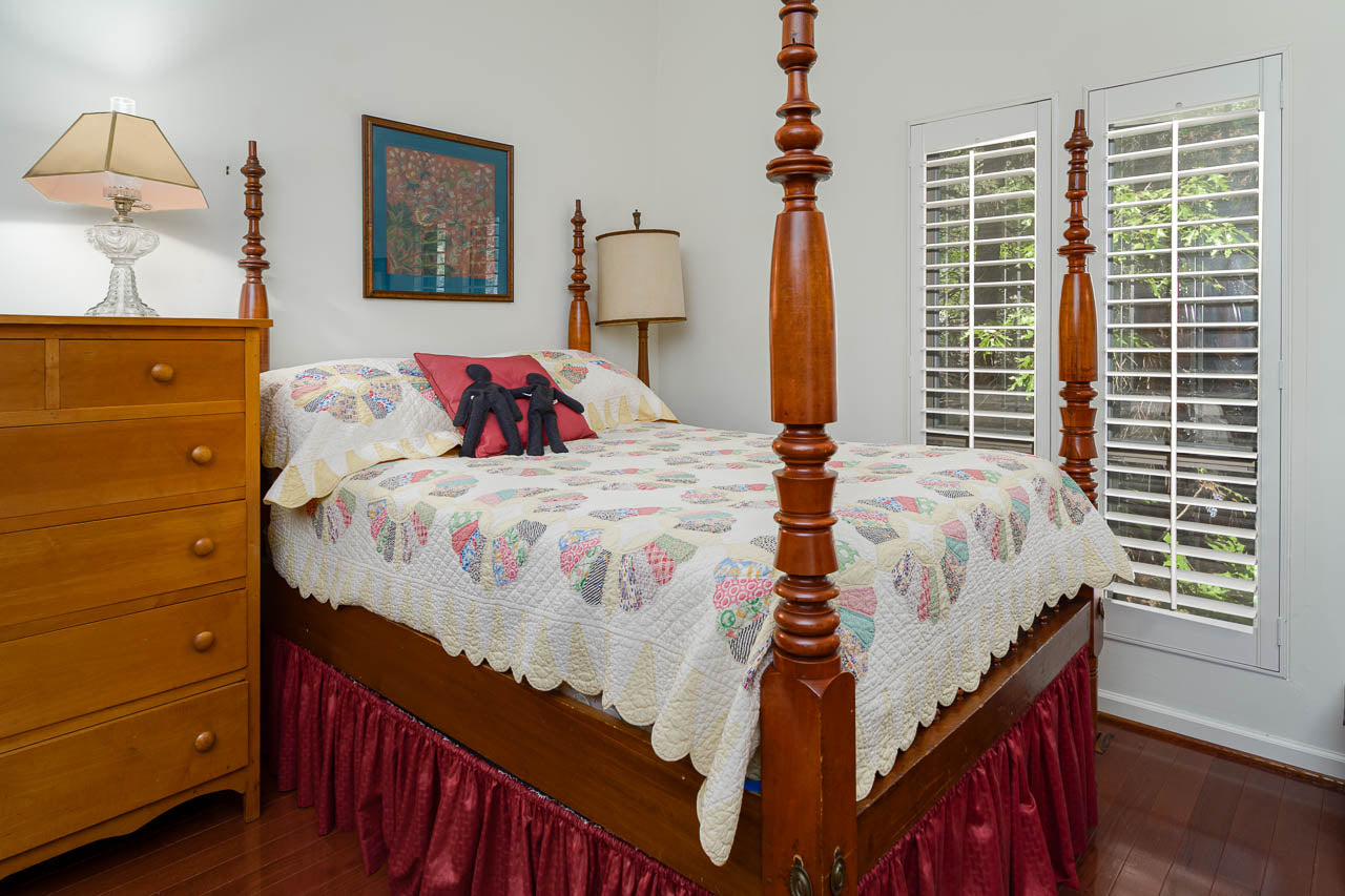 A cozy bedroom with a wooden four-poster bed, colorful patchwork quilt, red bed skirt, and decorative pillows. A wooden dresser, lamp, wall art, and shuttered windows complete the room.