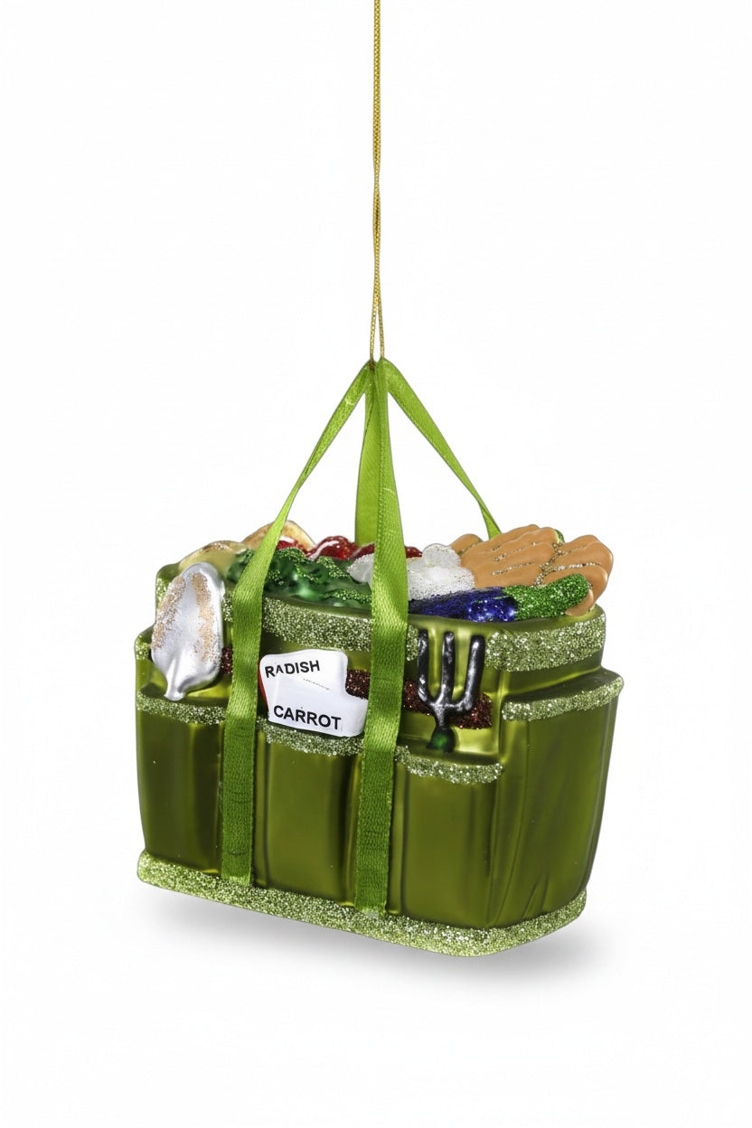 A 4-inch GARDENERS TOTE ORNAMENT by CODY FOSTER, shaped like a glass garden tote filled with gardening tools and seed packets labeled “Radish” and “Carrot.”.