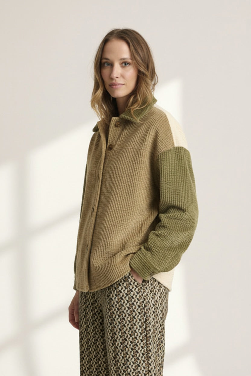 A woman with light brown hair wears the STORIATIPIC Corduroy Multi Shacket by STORIATIPIC—a tan and olive green button-up jacket crafted from soft cotton corduroy with a single chest pocket—paired with patterned pants. She stands against a plain light background, gazing at the camera with a neutral expression.
