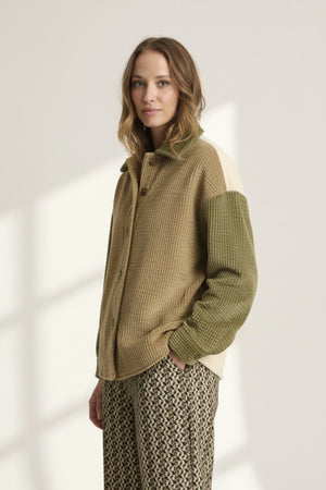 A woman with light brown hair wears the STORIATIPIC Corduroy Multi Shacket by STORIATIPIC—a tan and olive green button-up jacket crafted from soft cotton corduroy with a single chest pocket—paired with patterned pants. She stands against a plain light background, gazing at the camera with a neutral expression.