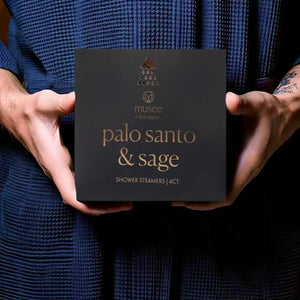 A box labeled “MUSEE - PALO SANTO & SAGE SHOWER STEAMERS” by MUSEE sits next to four white, round steamers featuring an elegant leaf design. The sleek black box with gold lettering is slightly open to reveal its illustrated inner packaging. This specialty MUSEE sage shower steamer elevates your daily routine, turning each shower into a luxurious aromatherapy experience.