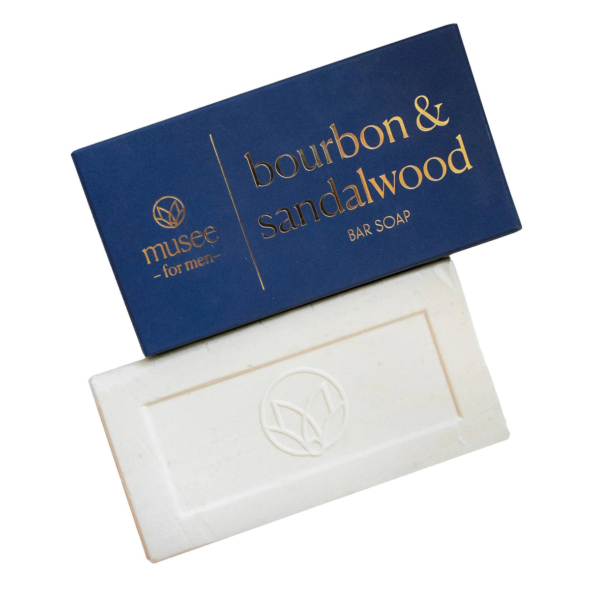 A rectangular white bar of MUSEE - BOURBON & SANDALWOOD BAR SOAP, known for its moisturizing properties, rests partially atop a blue box labeled with the product name and brand “MUSEE,” featuring a circular logo on both the soap and the packaging.