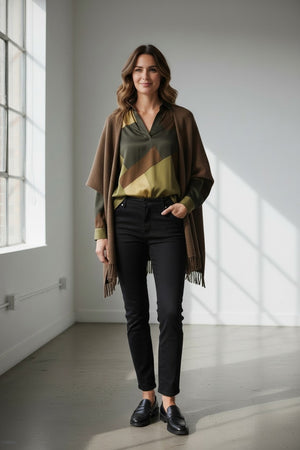 A woman stands indoors by a large window, sunlight streaming in and casting soft shadows on the floor, wearing the HARTFORD - VIRGINIE WOOL CAPE — a chic and casual brown cape from HARTFORD crafted from double-faced wool — layered over a patterned blouse, black pants, and black loafers.
