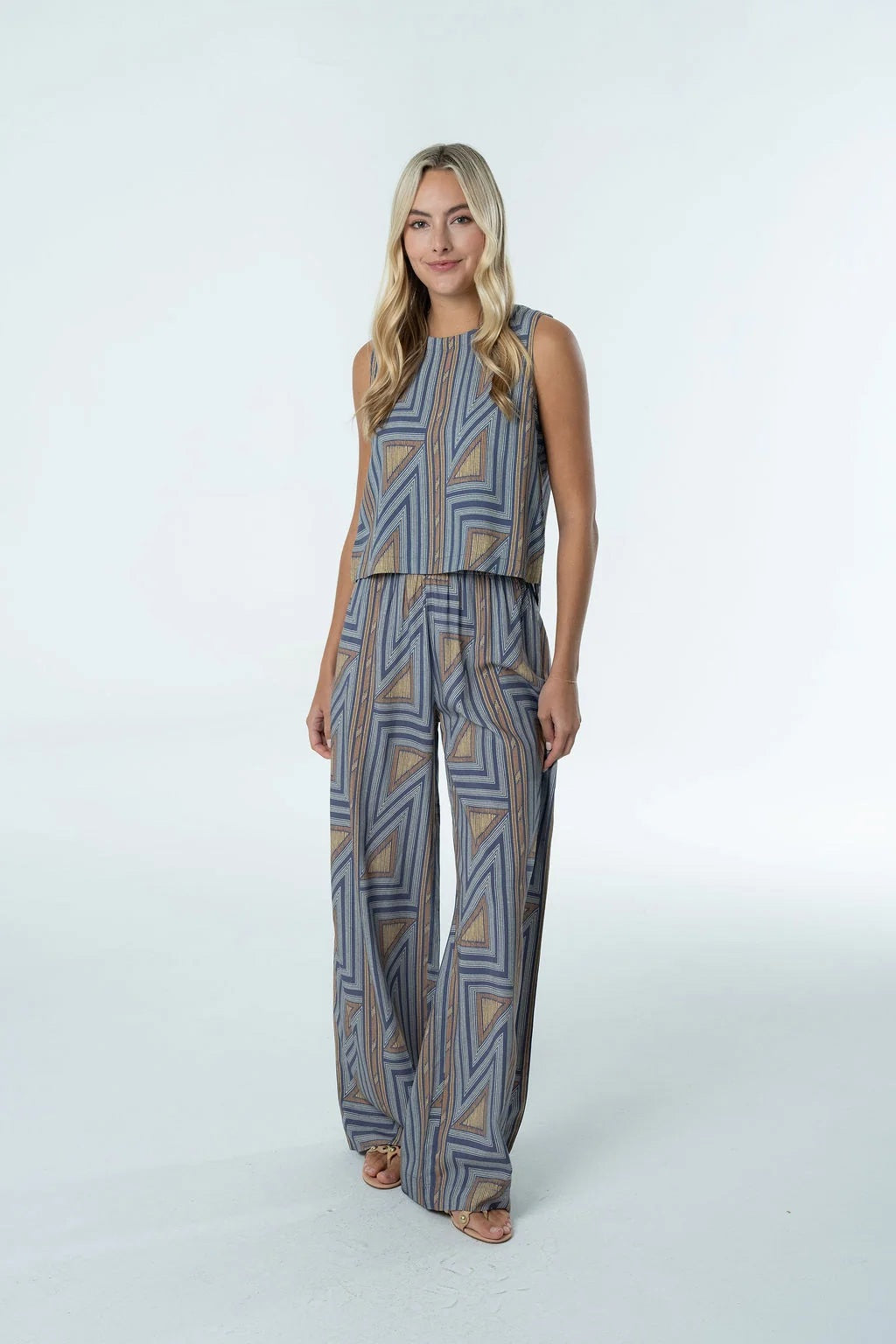 A woman stands against a plain white background wearing the BRITON COURT SARA PANT by BRITON COURT, a sleeveless jumpsuit with wide-leg pants featuring a blue and tan geometric pattern.