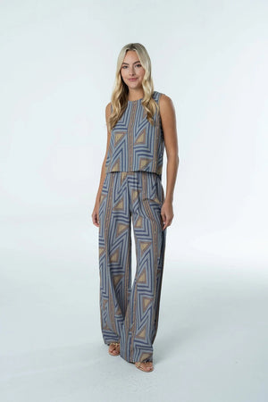 A woman stands against a plain white background wearing the BRITON COURT SARA PANT by BRITON COURT, a sleeveless jumpsuit with wide-leg pants featuring a blue and tan geometric pattern.