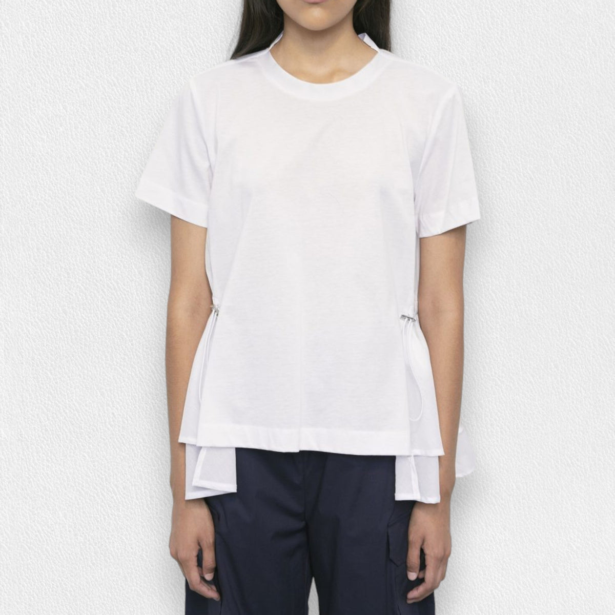 A person is standing against a light textured background wearing the MEIMEIJ - SIDE CINCH TOP, a white short-sleeve, round neck top with side slits and drawstrings. This flared T-shirt features a layered hem and is paired with navy blue pants. Their face is not fully visible.