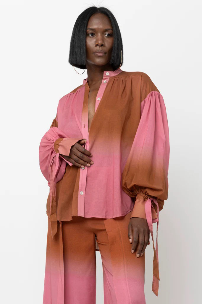 A person stands confidently wearing a stylish outfit consisting of the HOPE FOR FLOWERS Wrist Tie Blouse and matching pants in a gradient pink to brown color. Made from sustainable fabric, the blouse features balloon sleeves and a relaxed collar. The person has a serious expression and shoulder-length straight hair.