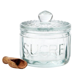 The ETUHOME Bistro Glass Sugar Cellar with Scoop by ETUHOME holds brown sugar on a white countertop near a folded white towel with red stripes and a wooden tray, adding classic charm to your kitchen storage collection.