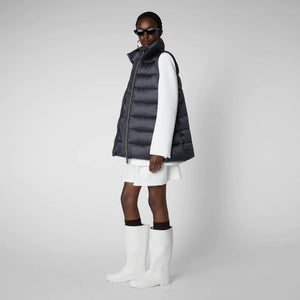 A person wearing a black cap, sunglasses, the SAVE THE DUCK - CORAL PUFFER VEST with animal-free down from SAVE THE DUCK over a black long-sleeve top, beige wide-legged pants, and black ankle boots stands against a plain background. The outfit appears to be styled for colder weather.