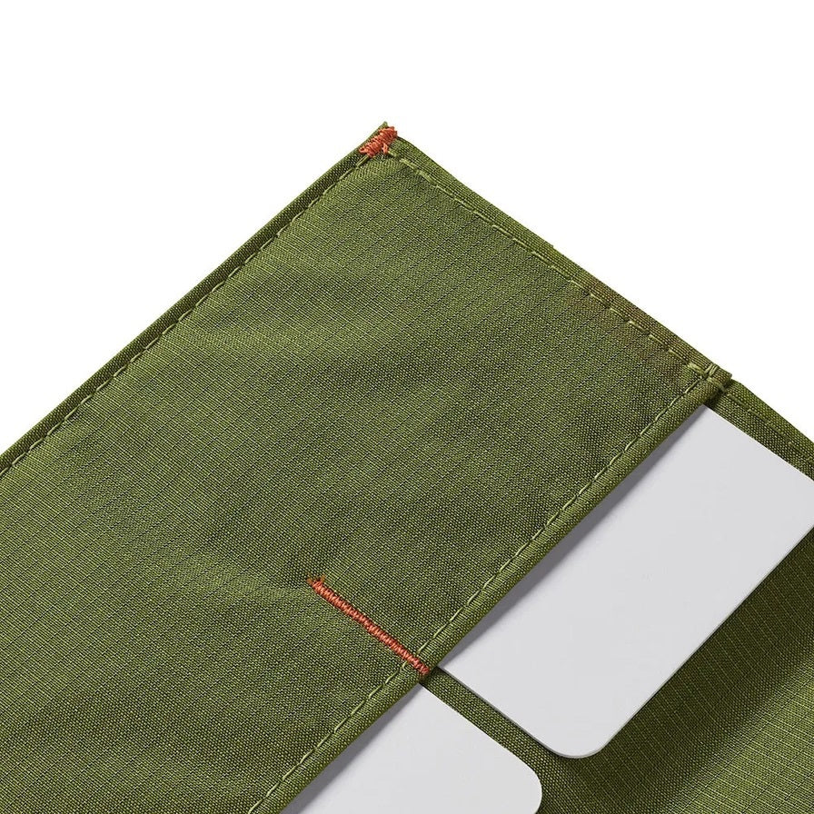 A Cala Green ALLETT RFID-blocking nylon wallet rests on a wooden surface beside a green notebook, portable stove, headlamp, black bag, pocket knife, camping gear, and round dark objects.