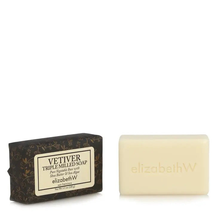 A rectangular bar of soap, enriched with shea butter, is placed next to its packaging. The cream-colored soap is inscribed with "elizabethW." The black packaging features gold floral designs and labels like "VETIVER TRIPLE MILLED SOAP 3.5OZ," along with other text in white and gold fonts, showcasing the brand name "E WIGHTMAN &amp; CO.
