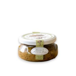 A jar of BELLA CUCINA - PORCINI & PARMESAN PESTO is displayed against a light gray background. The jar is topped with a white lid and adorned with a green label featuring an illustration of porcini mushrooms, alongside oval text labels indicating the product name and net weight of 6 oz (168 g).