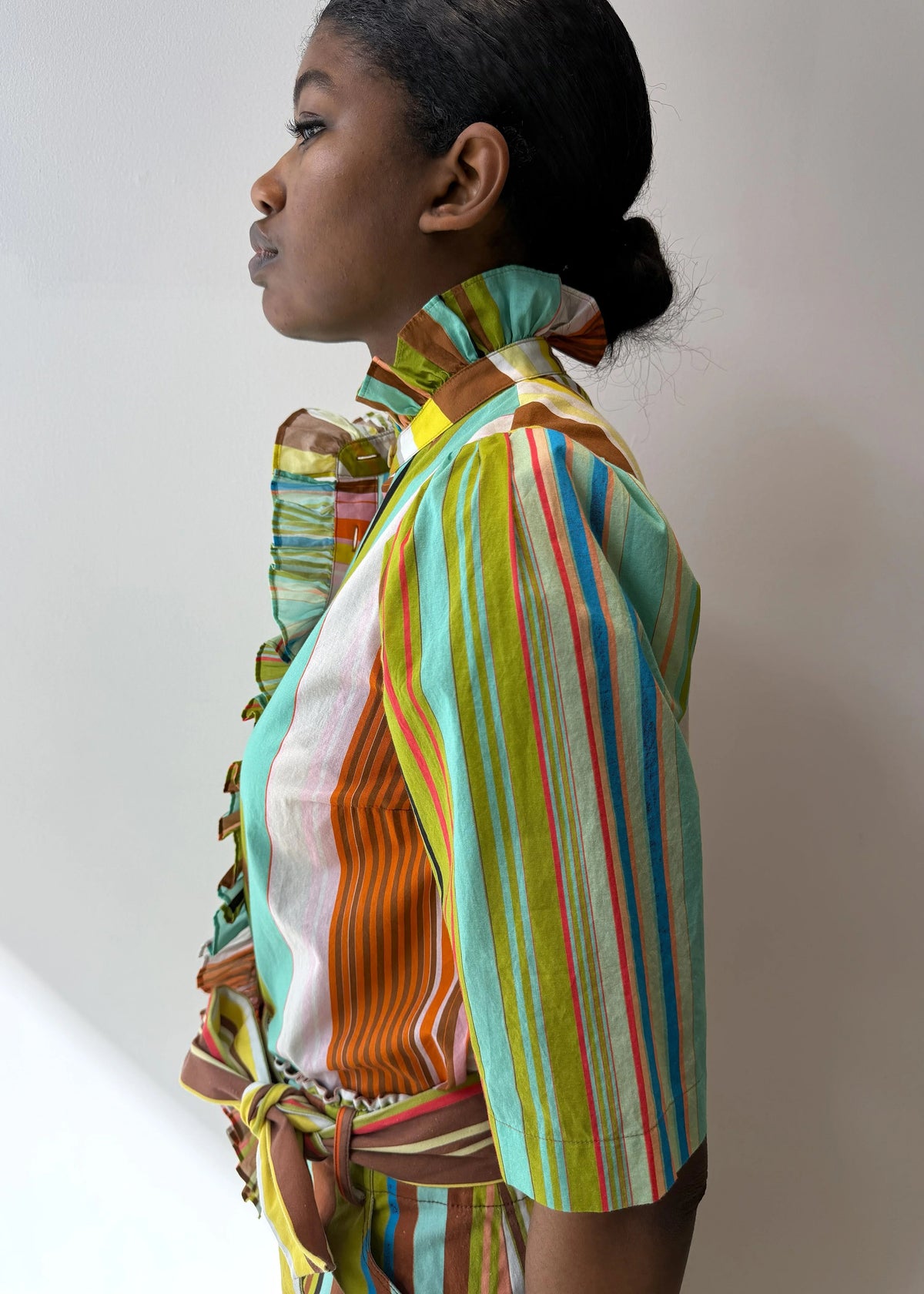 A person models the HOPE FOR FLOWERS Frilled Button Down Shirt in Combo Stripes by HOPE FOR FLOWERS, featuring vibrant vertical stripes and a ruffled collar, crafted from GOTS certified organic cotton for sustainability.