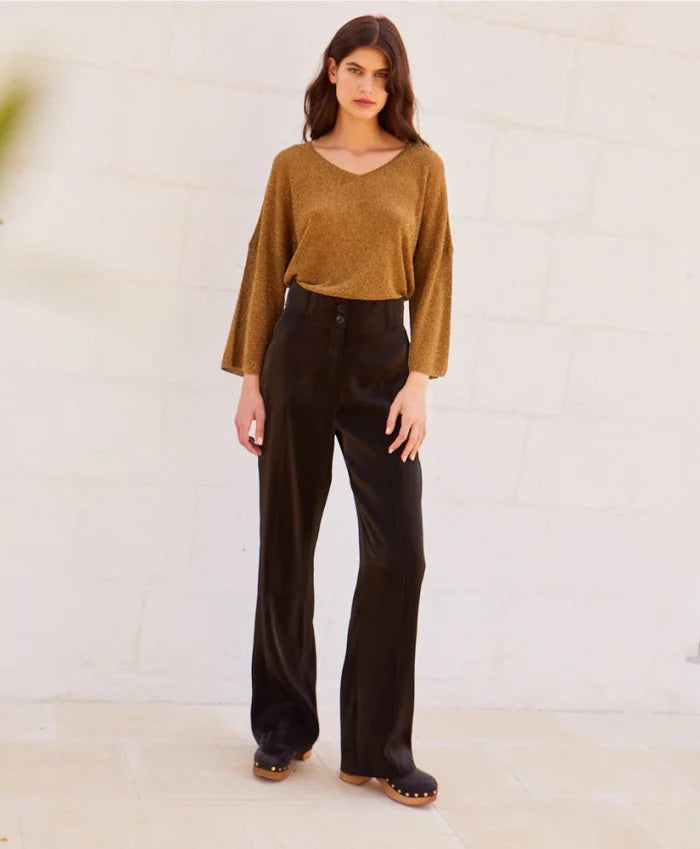 A person stands against a light-colored stone wall, wearing a loose-fitting, long-sleeved brown top with bio-galalite buttons paired with the high-waisted, wide-leg MANHATTAN PANT from MOMONI. They have long hair and are looking directly at the camera. The background is simple and uncluttered.