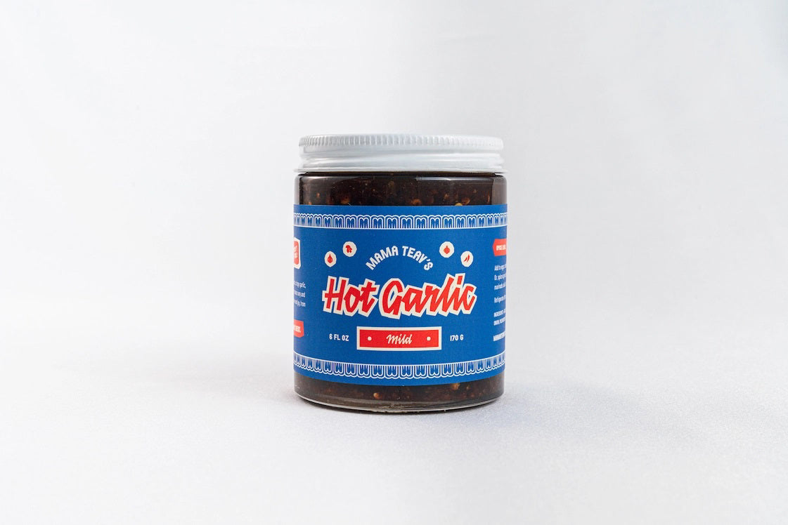 A jar of MAMA TEAV'S- MILD HOT GARLIC CHILI CRISPS is displayed against a plain white background. The jar features a blue label with the brand name "MAMA TEAV'S" prominently in the center and the word "MILD" beneath it, describing the product inside. Made with natural ingredients, this gluten-free Hot Garlic Chili Crisp is perfect for any meal.