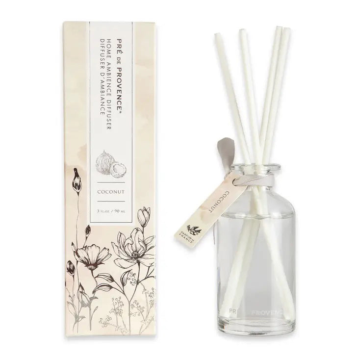 A glass reed diffuser bottle with white reeds is placed next to its packaging. The packaging, adorned with floral illustrations and the text &quot;HERITAGE REED DIFFUSER - COCONUT,&quot; showcases the quality products made by EUROPEAN SOAPS, LLC. A small tag reading &quot;Coconut&quot; is attached to the bottle.