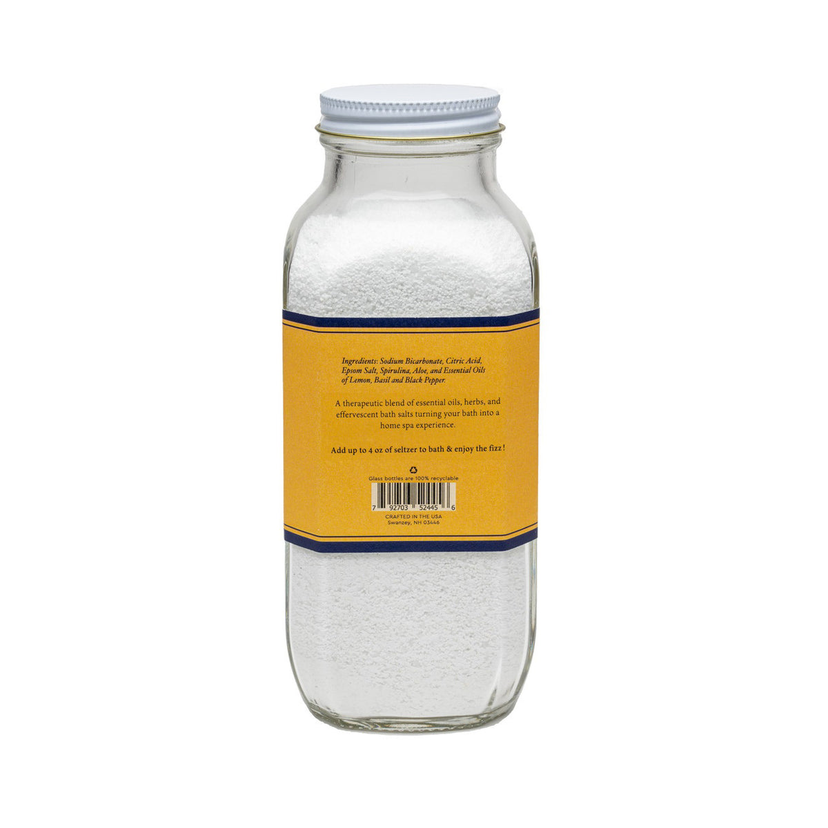 A clear glass jar with a white lid contains white bath salts. The jar&#39;s yellow and blue label reads: &quot;BAUDELAIRE - DETOX BATH SELTZER 18 OZ.&quot; Additional text indicates it is handmade, therapeutic, all-natural with essential oils and natural ingredients, featuring a proprietary formula.