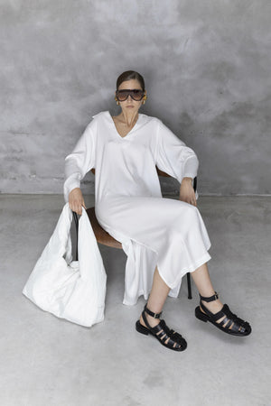 A woman in a long white dress sits on a chair against a gray wall, wearing large sunglasses, chunky black sandals, and gold earrings, holding a large white bag on her lap.