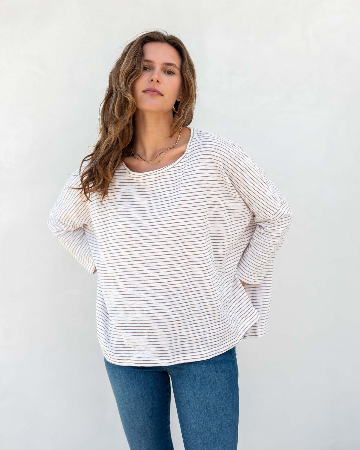 A woman with wavy brown hair models the MERSEA - CATALINA SLUB TEE—an oversized, long-sleeved white top with thin gray stripes—by MERSEA. She pairs it with blue jeans and stands against a plain, light background, hands behind her back.
