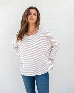 A woman with wavy brown hair models the MERSEA - CATALINA SLUB TEE—an oversized, long-sleeved white top with thin gray stripes—by MERSEA. She pairs it with blue jeans and stands against a plain, light background, hands behind her back.