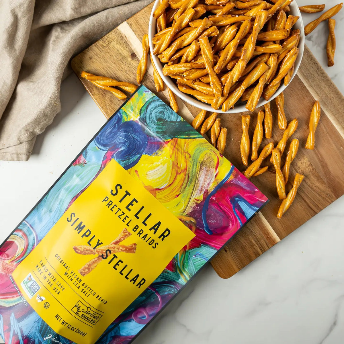 A vibrant bag of STELLAR SNACKS - SIMPLY STELLAR PRETZEL BRAIDS 5 OZ rests beside a white bowl filled with pretzel braids, set on a wooden board with a beige cloth nearby, all arranged on a white surface.