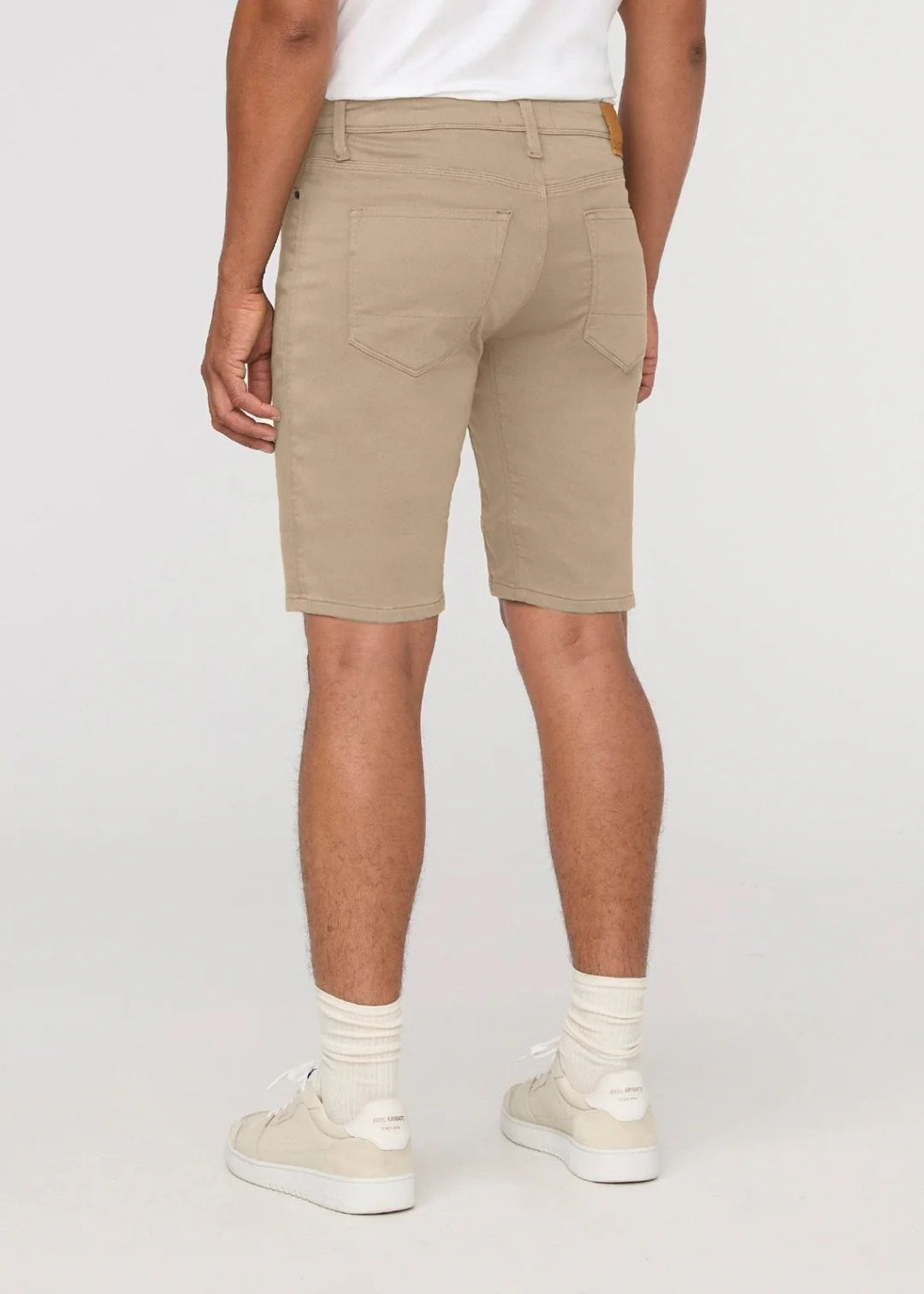 A person wearing a white t-shirt, DUER No Sweat Relaxed Shorts in Desert Khaki, white crew socks, white sneakers, and a wristwatch stands against a plain light background.