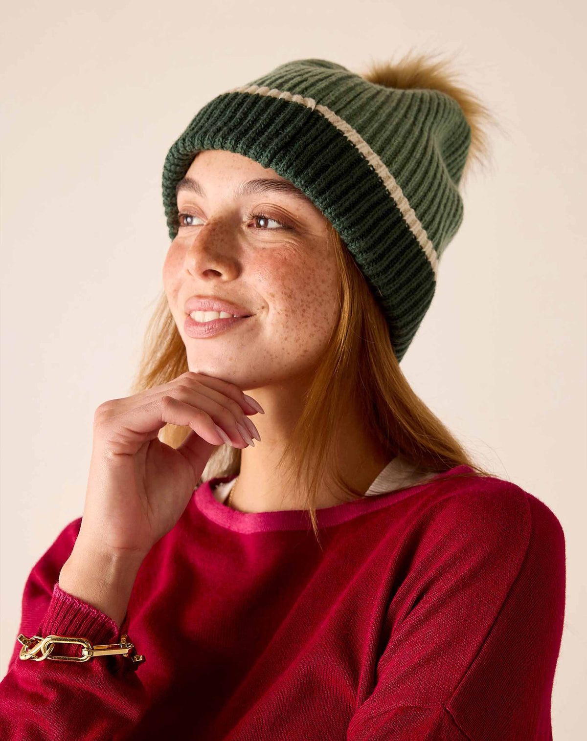The MERSEA Montauk Colorblock Beanie in Eden showcases a green ribbed knit design with a cream stripe and dark green edge, finished with a large brown faux fur pom-pom—an ideal cold-weather accessory from MERSEA, presented on a plain white background.