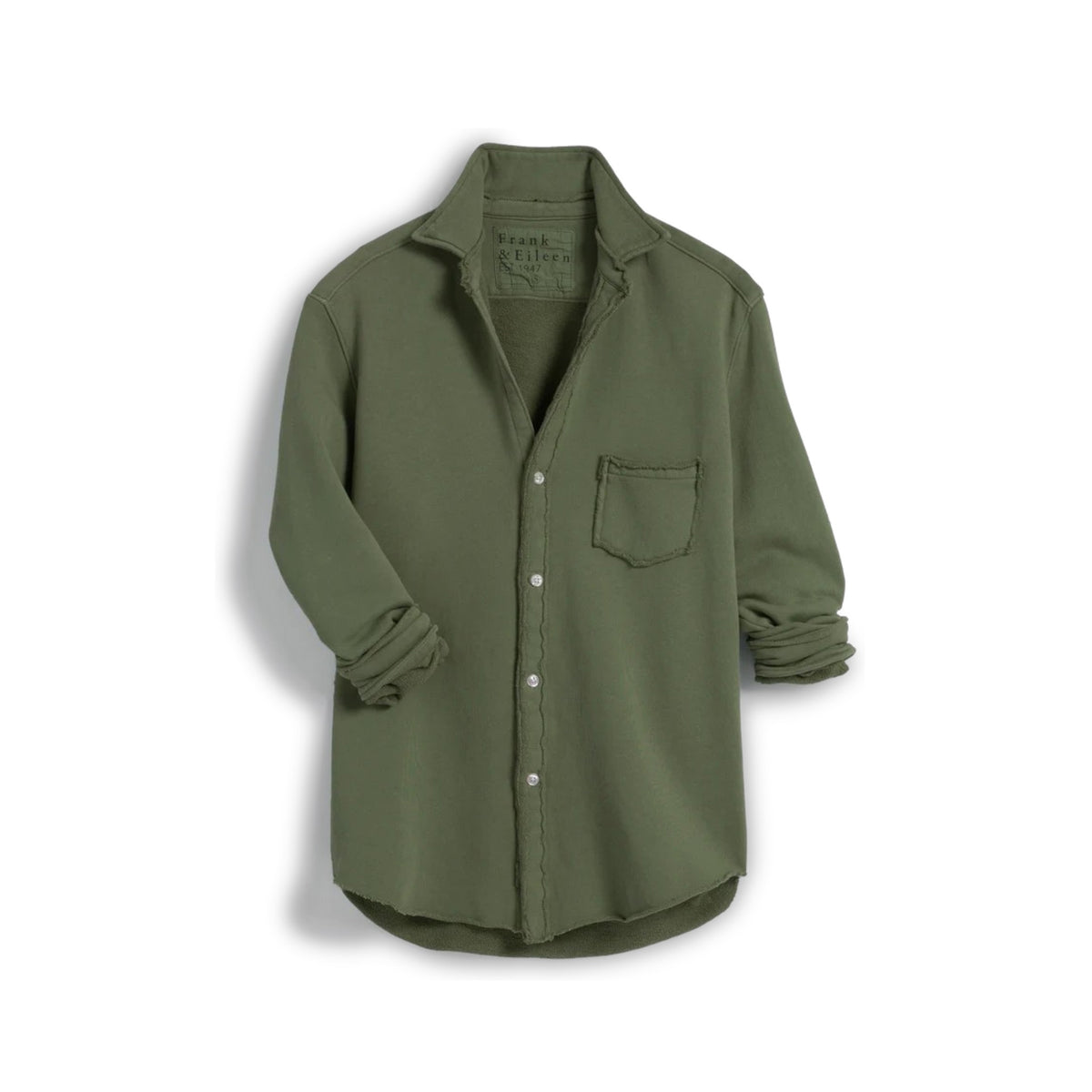 In a sunny, rustic kitchen, a woman smiles while wearing the FRANK &amp; EILEEN EILEEN Relaxed Button Up in Triple Fleece Caper, paired with black wide-leg pants and sandals. She touches her hair as sunlight fills the cozy space.