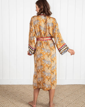 A woman stands barefoot by a dark fireplace, holding a drink, wearing the PRINTFRESH - BAGHEERA ECO SATIN ROBE IN TOBACCO from PRINTFRESH. Books and pampas grass adorn the mantel, while her reflection appears in the mirror behind her.