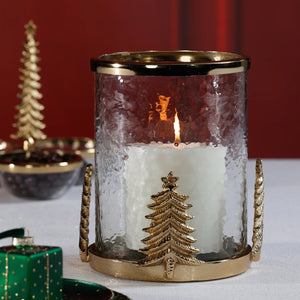 ZODAX GLASS AND ALLOY HURRICAN IN GOLD featuring a clear cylindrical glass and steel hurricane lamp with a gold-toned base, rim, and decorative gold Christmas tree embellishments on the sides.