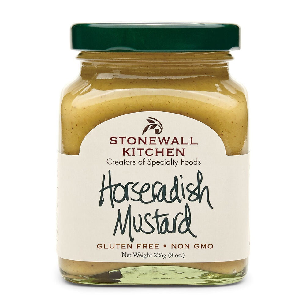 A ham, cheese, lettuce, and tomato sandwich made with STONEWALL KITCHEN - HORSERADISH MUSTARD sits beside an open jar of STONEWALL KITCHEN Horseradish Mustard, with mustard spread on the table. Chips and a drink appear blurred in the background.