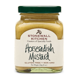 A ham, cheese, lettuce, and tomato sandwich made with STONEWALL KITCHEN - HORSERADISH MUSTARD sits beside an open jar of STONEWALL KITCHEN Horseradish Mustard, with mustard spread on the table. Chips and a drink appear blurred in the background.