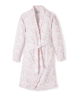 The PETITE PLUME - DORSET FLORAL ROBE by PETITE PLUME, displayed hanging, features a subtle, all-over pink floral pattern on a white background. This elegant robe is made from 100% cotton and includes long sleeves, a shawl collar, and a fabric belt tied at the waist—perfect for luxurious sleepwear.