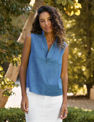 A woman with curly hair wears the FRANK & EILEEN Olivia Popover Henley Tank in Famous Denim Stonewashed Indigo, paired with a white skirt, standing outdoors in a sunlit garden surrounded by trees.