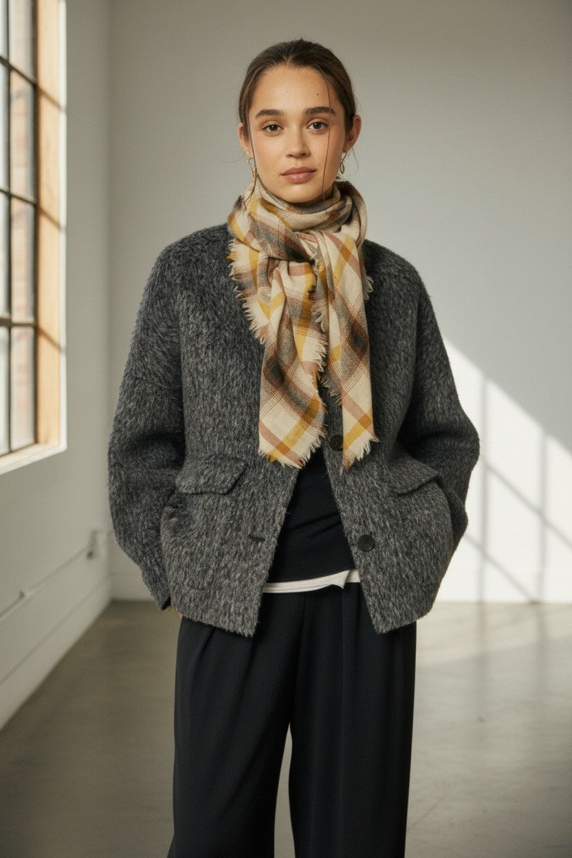 A person stands indoors with their hands in their jacket pockets, wearing a gray textured jacket, black pants, and the HARTFORD Checks Wool Scarf in Ecru and Butter by HARTFORD. Sunlight streams through large windows in the background.