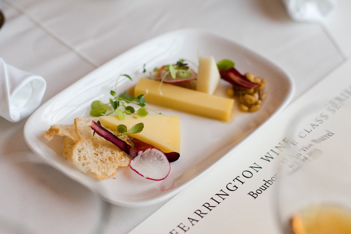 A rectangular white plate with slices of cheese, microgreens, thinly sliced radish, crispy bread, and pickles, arranged neatly on a white tablecloth with a printed menu partially visible underneath.