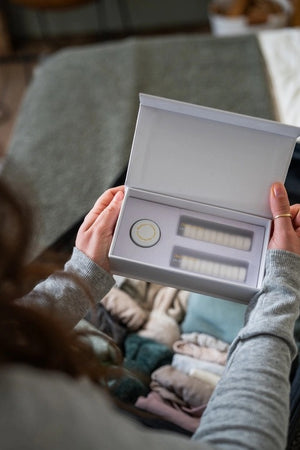A person in a gray sweater and jeans packs folded clothes and reusable scented towels into a suitcase while holding the MADISON GREEN “REST YOUR EYES ON THE PLANE” white rectangular box. A wooden dresser and a small plant can be seen in the background.