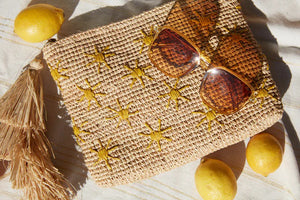 The MAR Y SOL - SOLEIL CLUTCH by MAR Y SOL, featuring yellow sun embroidery and an oversized tassel, rests on a light cloth with three lemons and brown sunglasses. Sunlight casts playful shadows across this stylish display.