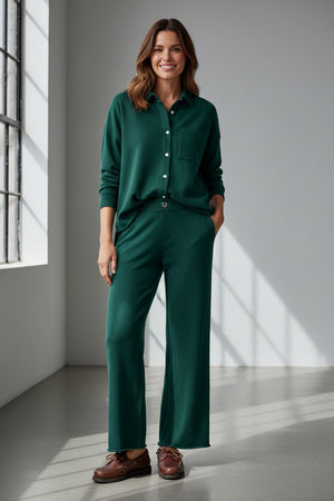 A woman stands indoors with her hands in her pockets, wearing the FRANK & EILEEN Riley Cropped Trouser Pant in Triple Fleece Pine from the California Heritage Collection, paired with a matching dark green button-up shirt and brown shoes, as sunlight streams through a large window.