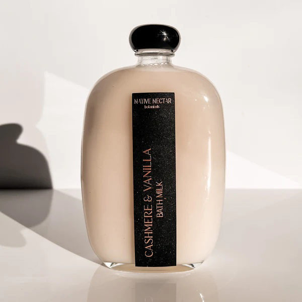 A clear glass bottle of NATIVE NECTAR’s CASHMERE &amp; VANILLA BATH MILK sits on a white surface with soft shadows—an ideal choice for silky, smooth skin.