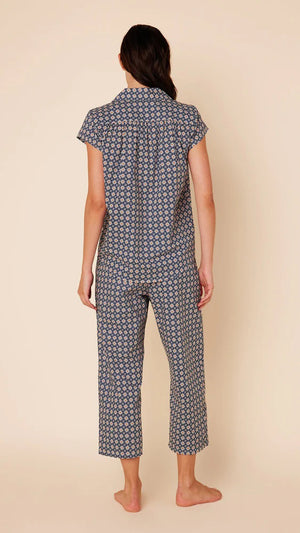 A woman stands barefoot against a plain beige background, wearing the CAT'S PAJAMAS - NEWPORT DAISY LUXE PIMA COTTON CAPRIS PAJAMA from CAT'S PAJAMAS. The short-sleeved button-up top and matching loose-fitting capri pants feature a small, intricate pattern in shades of blue and white, crafted from 100% Pima cotton.