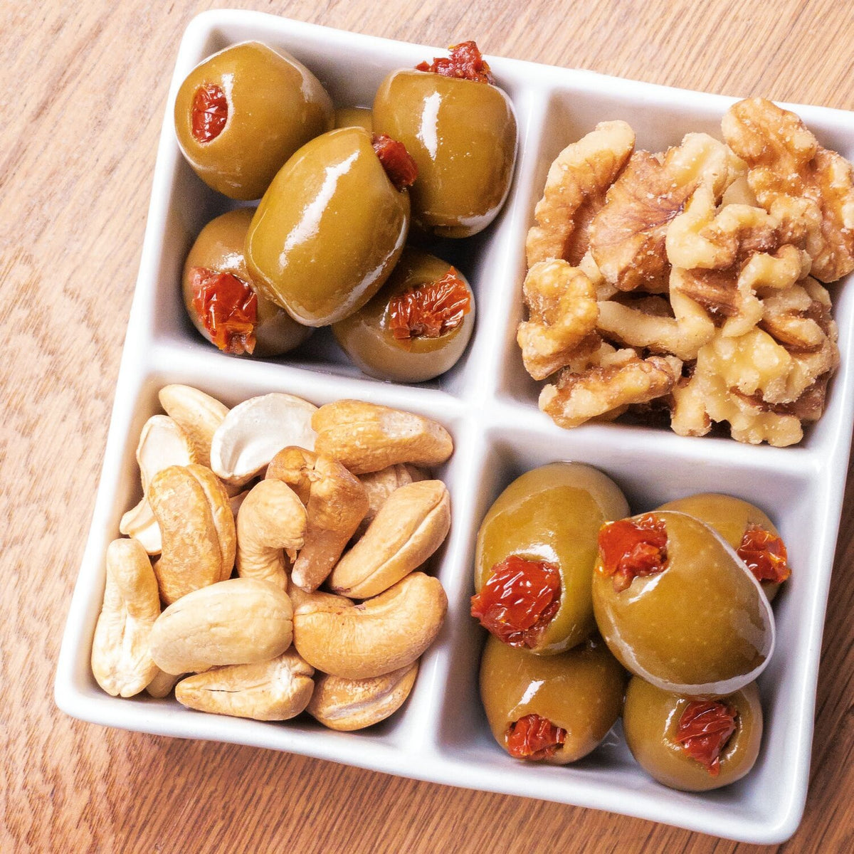 A square white dish with four sections holds DIVINA - SUNDRIED TOMATO STUFFED OLIVES, walnuts, and cashews, arranged on a wooden surface. These delicious olives by DIVINA are made in Greece and provide an authentic Mediterranean taste experience.