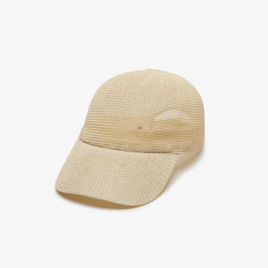 A woman with long, wavy blonde hair wears the WYETH HATS - PAULA MESH CAP IN NATURAL by WYETH and a white tank top, sitting against a plain light background and looking to the left with a neutral expression.