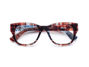 A person wearing CADDIS - MIKLOS READERS IN MADRAS PATCHWORK glasses with thick-rimmed, multicolored patterned frames against a solid red background.