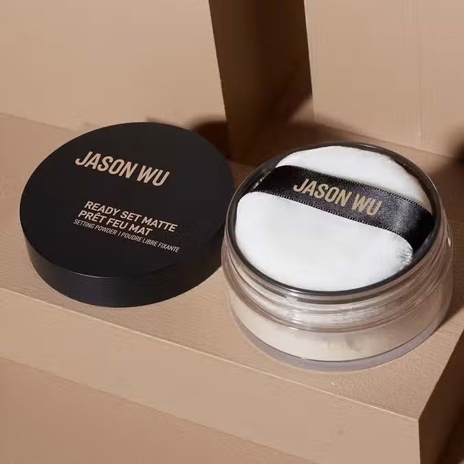 A container of JASON WU BEAUTY - READY SET MATTE 1 - TRANSLUCENT BANANA setting powder sits on a light brown textured surface. The cylindrical container has a black lid with the brand name in white capital letters and product details in both English and French, ensuring a flawless matte finish for any makeup look.