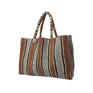 Wearing blue jeans and light espadrilles, the person carries the GABRIELE FRANTZEN LITTLE SISTER WEEKENDER BAG TOBACCO in earthy brown, beige, and gray stripes. Their white shirt is partially visible.