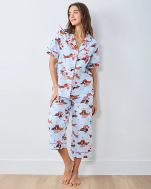 A woman with long, wavy brown hair stands barefoot against a white wall, wearing the PRINTFRESH Dachshund Date Short Sleeve Top & Cropped Pants Pajama Set in Blue. The organic cotton set from PRINTFRESH features short sleeves and cropped pants adorned with a charming dachshund print. She smiles slightly.