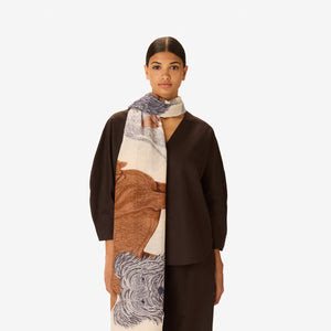 A woman in a dark outfit stands with her hands in her pockets, draped in the INOUI EDITIONS - TOUTOU Scarf in White by INOUI, featuring intricate dark brown and navy animal illustrations. She is set against a plain white background.