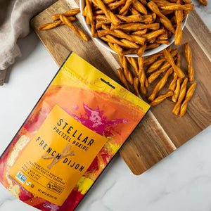 A vibrant bag in shades of orange and yellow—labeled “STELLAR SNACKS FRENCH DIJON PRETZEL BRAIDS”—features a vegan butter braid with honey mustard style seasoning. The packaging highlights that the product is Non-GMO, and “Baked with Love” is prominently displayed at the bottom.
